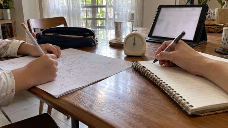 Two hands, a child's and a parent's, rest over a worksheet, notebook, kitchen timer and glass of water on a dining table.
